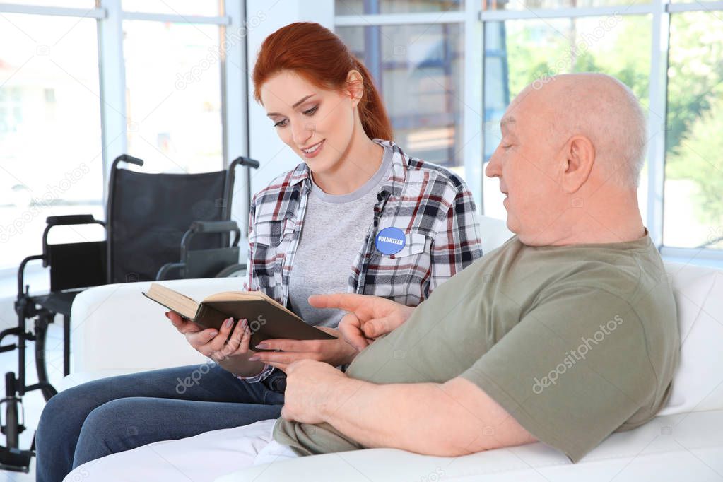 Young female volunteer reading book to senior man in light room