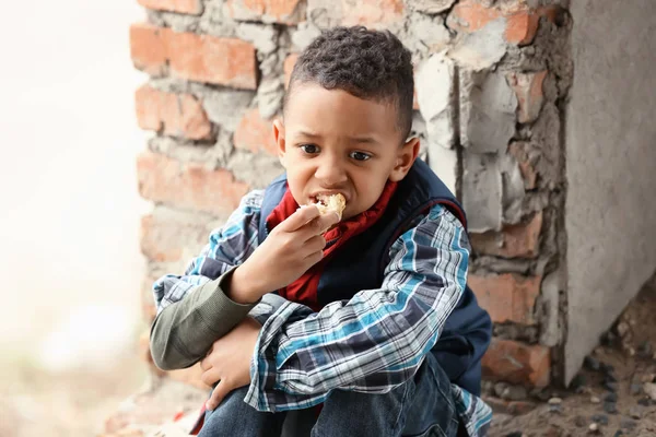 Aranyos kisfiú ül darab kenyér elhagyott épület emeletén. A szegénység fogalma — Stock Fotó