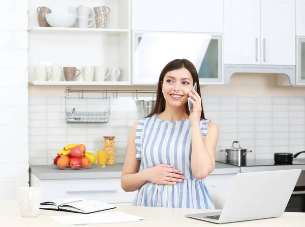 Young beautiful pregnant woman talking by phone while working in kitchen — Stock Photo, Image