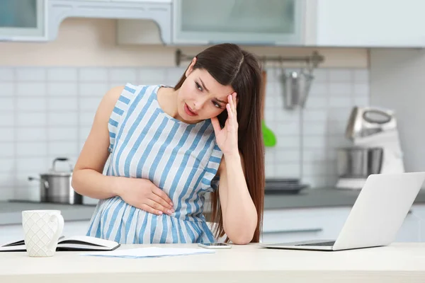 Krásná mladá těhotná žena trpí bolesti hlavy při práci s přenosným počítačem v kuchyni — Stock fotografie