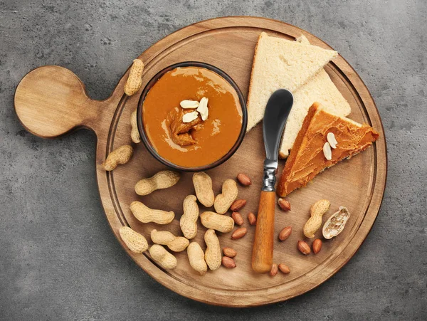 Placa de madeira com deliciosa manteiga de amendoim, torradas e nozes na mesa — Fotografia de Stock