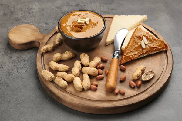 Wooden board with delicious peanut butter, toasts and nuts on table — Stock Photo, Image