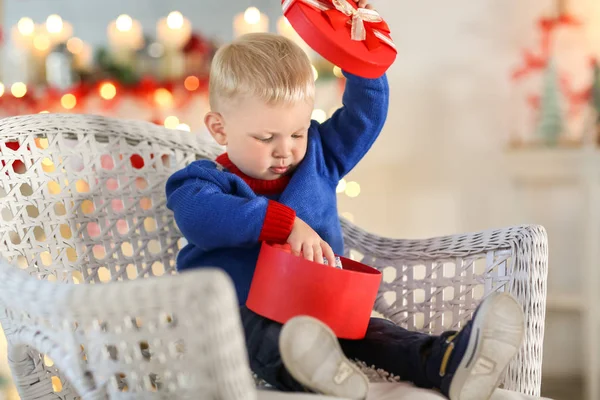 Симпатичный маленький мальчик с подарочной коробкой сидит в плетеном кресле на размытом фоне — стоковое фото