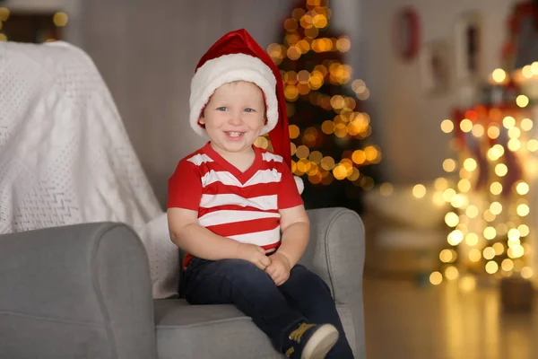 可爱的小男孩，在圣诞老人的帽子，坐在扶手椅上对模糊的圣诞灯 — 图库照片
