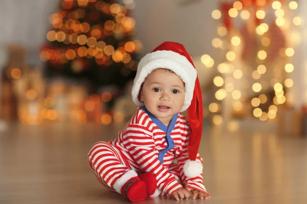 Söt liten baby med Tomtemössa på golvet och suddig julbelysning på bakgrund — Stockfoto