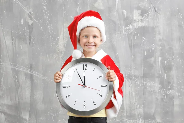 Linda niña en traje de Santa Claus con reloj sobre fondo grunge. Concepto de cuenta atrás de Navidad —  Fotos de Stock