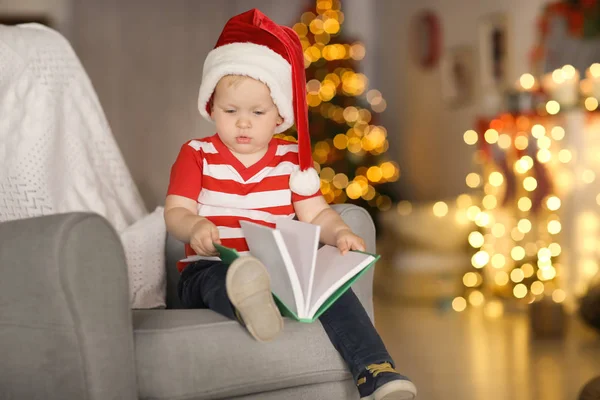 Netter kleiner Junge mit Weihnachtsmütze und Buch im Sessel gegen verschwommene Weihnachtsbeleuchtung — Stockfoto