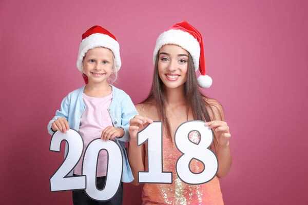 Young woman and cute little girl in Santa hats with paper figures 2018 on color background. Christmas celebration concept
