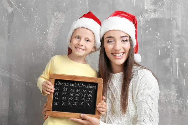 Молодая женщина и милая маленькая девочка в шляпах Санты с доской подсчета дней до Рождества, на гранж фоне — стоковое фото