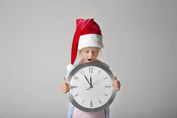 Linda niña en sombrero de Santa con reloj sobre fondo claro. Concepto de cuenta atrás de Navidad — Foto de Stock