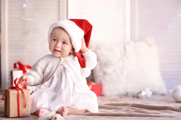 Mignon petit bébé avec chapeau de Père Noël et boîte cadeau assis sur le sol sur fond flou — Photo