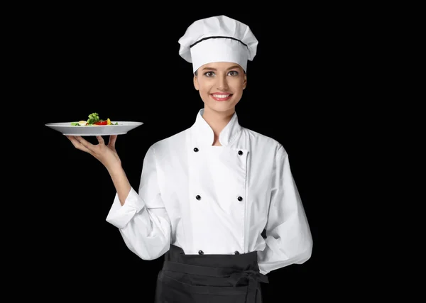 Jovem chef feminino segurando prato com salada no fundo escuro — Fotografia de Stock