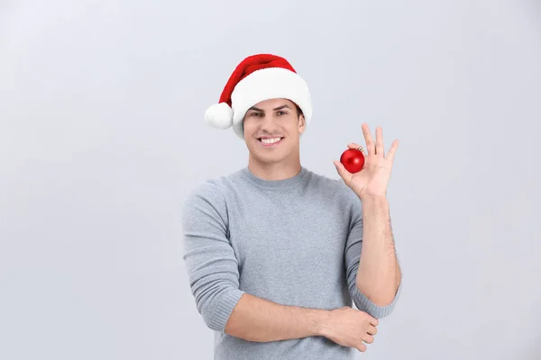 Bello uomo in cappello di Natale che tiene lucida bagattella su sfondo chiaro — Foto Stock