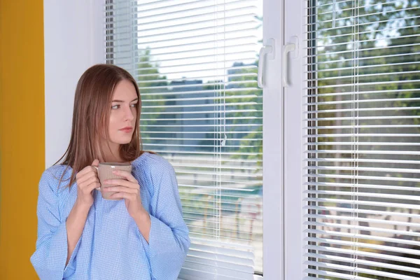 Belle jeune fille regardant dans la fenêtre et tenant une tasse de café ou de thé dans les mains — Photo