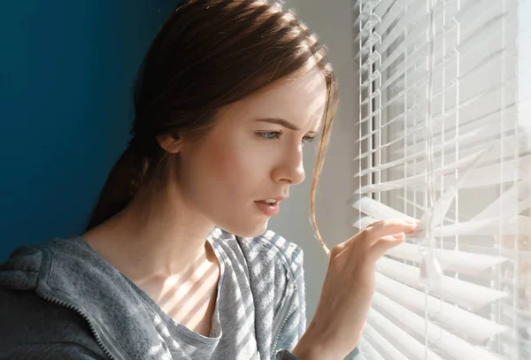 Belle jeune fille séparant lamelles de stores et regardant par la fenêtre — Photo