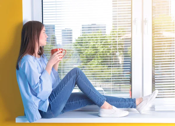 Hermosa chica joven mirando en la ventana y sosteniendo la taza de café o té en las manos — Foto de Stock