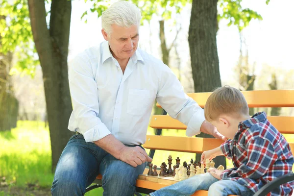 Lindo niño pequeño con su abuelo jugando ajedrez en el banco en el parque de primavera — Foto de Stock