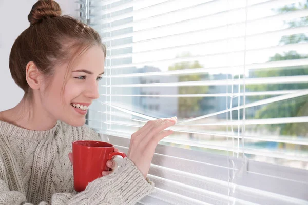 Belle jeune fille regardant dans la fenêtre et tenant une tasse de café ou de thé — Photo