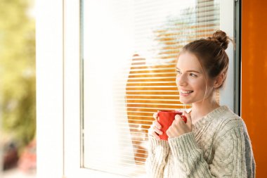 Beautiful young girl looking in window and holding cup of coffee or tea clipart