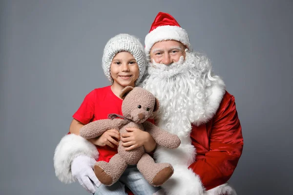 Niedlicher kleiner Junge mit Teddybär auf dem Schoß des Weihnachtsmannes vor farbigem Hintergrund — Stockfoto