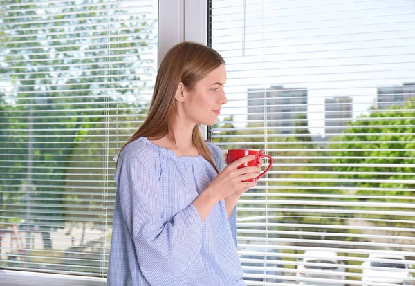 Belle jeune fille regardant dans la fenêtre et tenant une tasse de café ou de thé dans les mains — Photo