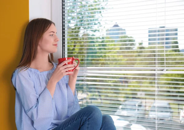 Belle jeune fille regardant dans la fenêtre et tenant une tasse de café ou de thé dans les mains — Photo