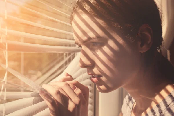 Hermosa chica joven separando listones de persianas y mirando a través de la ventana — Foto de Stock