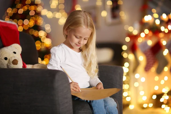 Sevimli kız koltukta oturan ve evde Noel Baba'ya mektup yazma — Stok fotoğraf