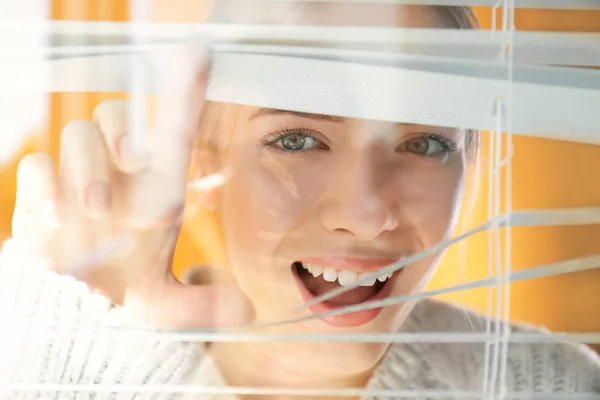 Beautiful young girl separating slats of blinds and looking through window — Stock Photo, Image