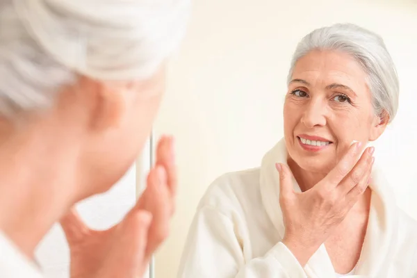 Beautiful elderly woman applying face cream in front of mirror at home — Stock Photo, Image