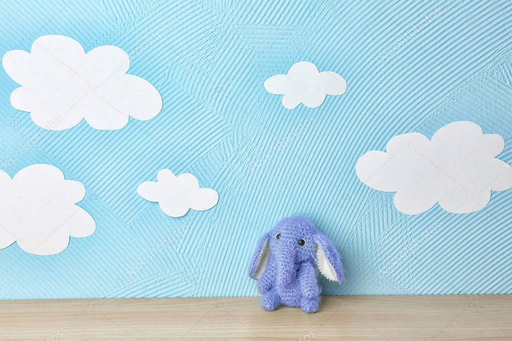 Cute knitted toy elephant on wooden table near blue wall with paper clouds