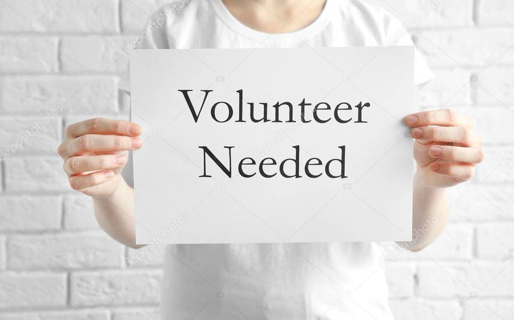 Woman holding sheet of paper with text VOLUNTEER NEEDED on light background