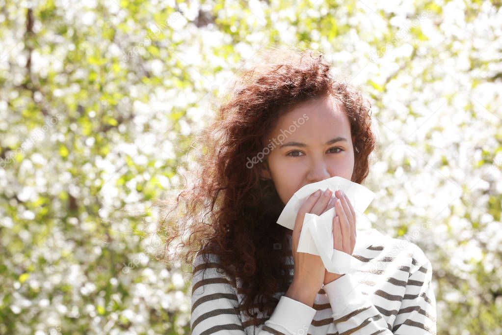 Allergy concept. Sneezing young girl with nose wiper among blooming trees in park
