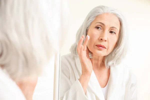 Belle femme âgée appliquant de la crème visage devant le miroir à la maison — Photo