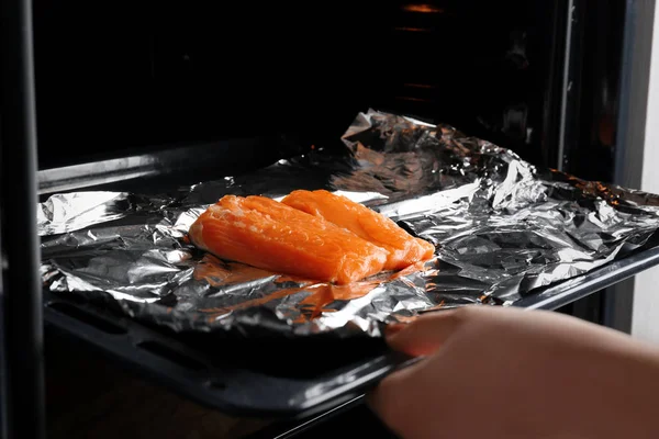 Женская рука кладет в духовку лист для выпечки с лососем на фольге — стоковое фото