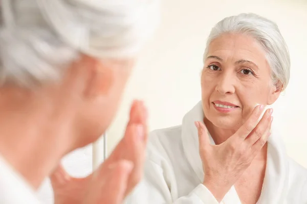 Beautiful elderly woman applying face cream in front of mirror at home — Stock Photo, Image