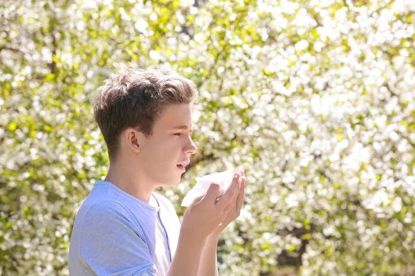 Allergy concept. Sneezing young boy with nose wiper among blooming trees in park — Stock Photo, Image