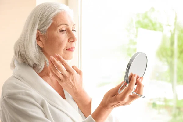 Wanita tua cantik memegang cermin dan menerapkan krim wajah di rumah — Stok Foto