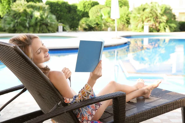 Όμορφη νεαρή γυναίκα ανάγνωση βιβλίο, ενώ χαλαρώνοντας στην ξαπλώστρα κοντά στην πισίνα — Φωτογραφία Αρχείου