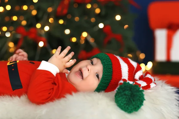 Cute little baby in Santa costume lying against blurred Christmas lights background — Stock Photo, Image