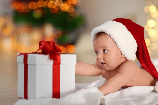 Schattige kleine baby in kerstmuts met een geschenkdoos liggend op de vloer tegen wazig Kerstverlichting — Stockfoto