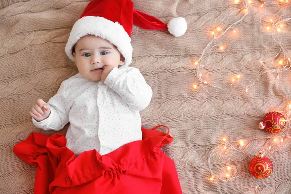 Lindo bebé con sombrero de Santa en bolsa roja y luces de Navidad en tela de punto —  Fotos de Stock