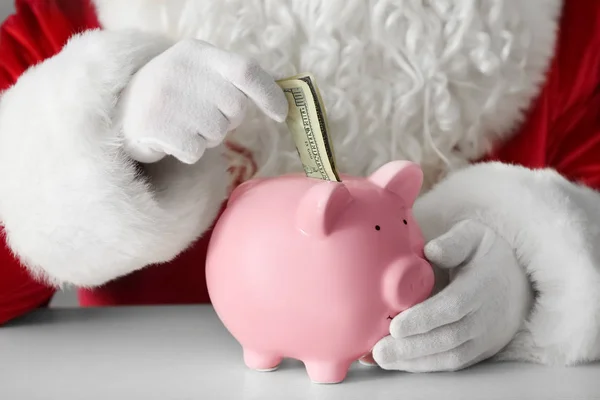 Weihnachtsmann legt Geld in Sparschwein am Tisch — Stockfoto