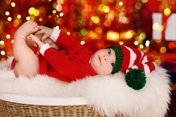 Niedliches kleines Baby im Weihnachtsmannkostüm vor verschwommenem Weihnachtsbeleuchtungshintergrund liegend — Stockfoto