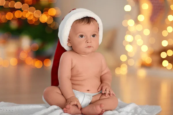 Söt liten baby i Santa hatt sitter på golvet mot suddig julbelysning — Stockfoto