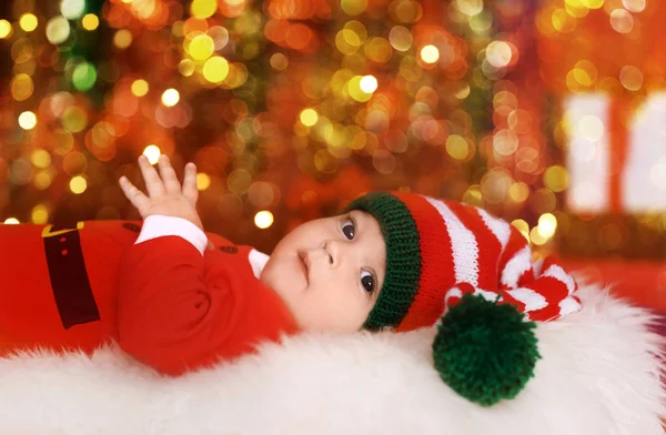 Niedliches kleines Baby im Weihnachtsmannkostüm vor verschwommenem Weihnachtsbeleuchtungshintergrund liegend — Stockfoto