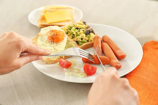 Woman eating tasty fried egg, sausages and toasts on table in kitchen