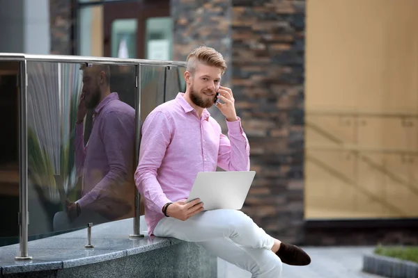 Молодой блоггер с ноутбуком разговаривает по смартфону, на открытом воздухе — стоковое фото
