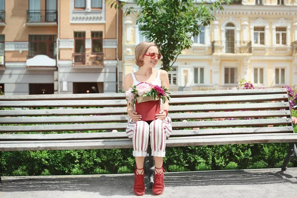 Menina bonita sentada no banco com flores de peônia na caixa de presente, ao ar livre — Fotografia de Stock