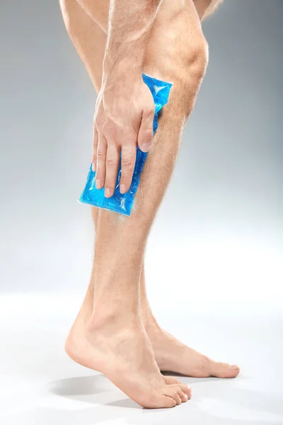 Giovane uomo che applica impacco freddo alla gamba su sfondo chiaro — Foto Stock